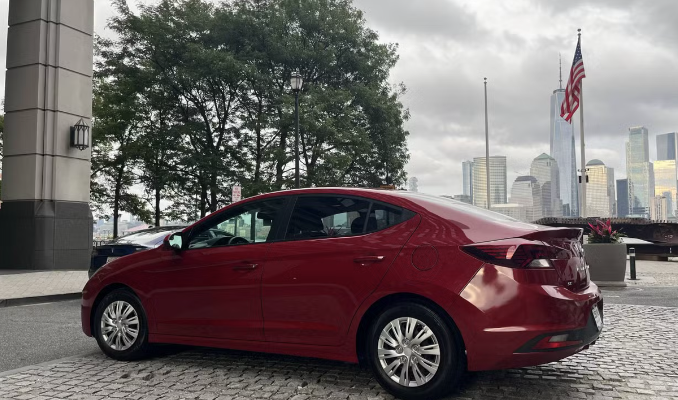 Red Hyundai Elantra parked near the Jersey City waterfront with the Manhattan skyline and an American flag in the background